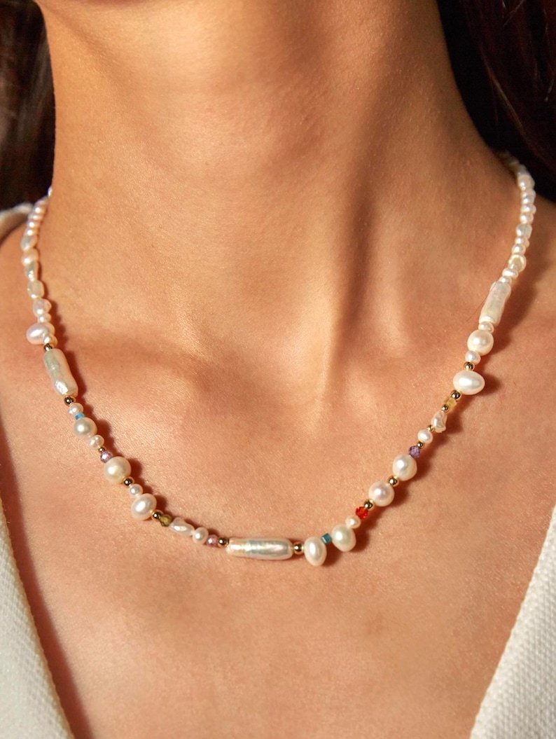 May include: A necklace featuring a mix of white pearls, small gold beads, and colourful accents. The necklace is made up of pearls of various shapes and sizes. The necklace is worn around the neck.