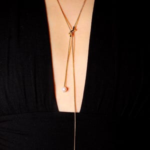 May include: A gold-toned lariat necklace featuring a thin chain tied in a central knot. The necklace ends in two distinct points, each finished with a single white pearl bead of different sizes, hanging at varying lengths against a black fabric.