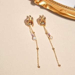 May include: A pair of gold-colored earrings with a textured, organic design. Each earring features a chain with small gold beads, a pearl, and small purple beads. The earrings are displayed on a light-colored surface.