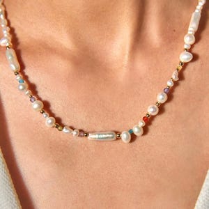 Op de afbeelding: Een ketting bestaande uit een streng witte zoetwaterparels in diverse vormen, waaronder langwerpige en ronde exemplaren, afgewisseld met kleine goudkleurige metalen kralen en piepkleine veelkleurige edelsteenaccenten over de gehele lengte van het sieraad.