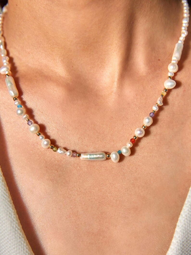 Op de afbeelding: Een ketting bestaande uit een streng witte zoetwaterparels in diverse vormen, waaronder langwerpige en ronde exemplaren, afgewisseld met kleine goudkleurige metalen kralen en piepkleine veelkleurige edelsteenaccenten over de gehele lengte van het sieraad.