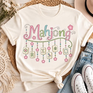 Può includere: Una t-shirt a maniche corte color crema con la parola Mahjong in lettere colorate e decorate. Sotto la scritta, pende una ghirlanda decorativa con fili di perline, fiori di ciliegio rosa e sei tessere del mahjong individuali.