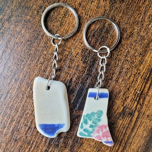 May include: Two keyrings with ceramic pendants. One pendant is off-white with a blue base, the other is off-white with a blue stripe and floral design in pink and green. Both have silver key rings and chains.