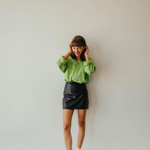 May include: A person wearing a bright green button-down shirt and a black leather mini skirt. The outfit is completed with black platform shoes and white socks. The background is a plain white wall.