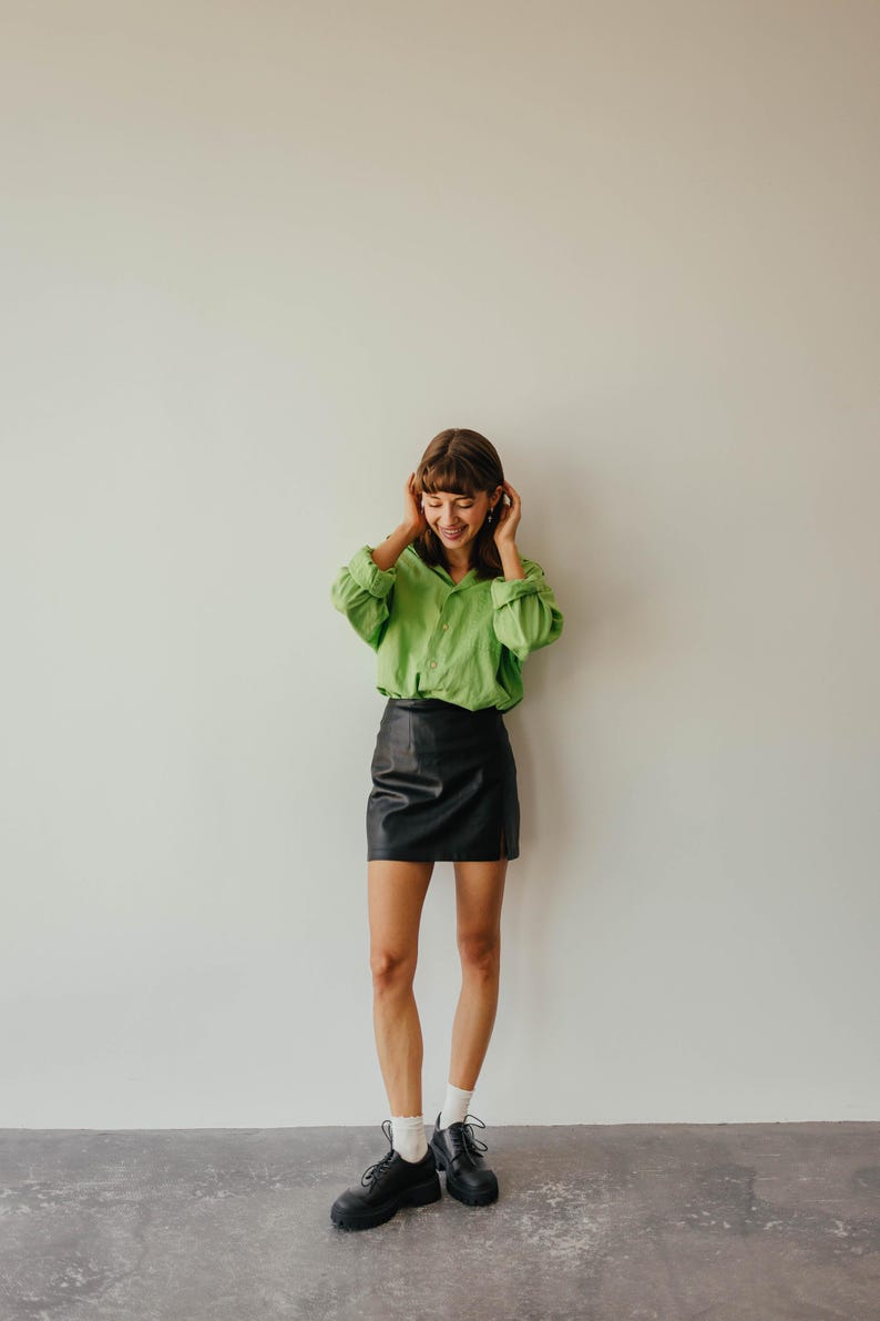 May include: A person wearing a bright green button-down shirt and a black leather mini skirt. The outfit is completed with black platform shoes and white socks. The background is a plain white wall.