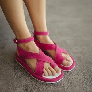 Peut inclure: Sandales rose vif avec des lanières croisées et des brides de cheville. Les sandales ont une semelle blanche et une fermeture à boucle. Les chaussures sont faites d'une matière douce et sont conçues pour le confort.