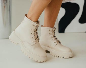 Puede incluir: Botines color crema con cordones y suelas gruesas y dentadas. Las botas tienen una puntera redondeada y un cuello acolchado. Los cordones son del mismo color que las botas. Las botas son de material liso.