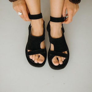 May include: Black suede sandals with ankle straps. The sandals have a flat sole and a simple design, featuring a strap across the toes and another over the top of the foot. The image shows the sandals from a top-down perspective.