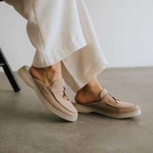 K&ouml;nnte beinhalten: Beige Wildleder-Slipper mit runder Spitze und kleinem goldfarbenen Quastendetail. Die Schuhe haben eine dicke, cremefarbene Sohle. Die Person tr&auml;gt eine wei&szlig;e Hose.