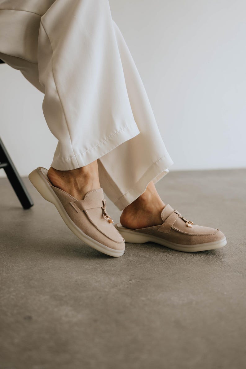 K&ouml;nnte beinhalten: Beige Wildleder-Slipper mit runder Spitze und kleinem goldfarbenen Quastendetail. Die Schuhe haben eine dicke, cremefarbene Sohle. Die Person tr&auml;gt eine wei&szlig;e Hose.