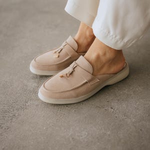 K&ouml;nnte beinhalten: Beige Wildleder-Slipper mit Quasten-Detail und einem kleinen goldfarbenen Akzent. Die Schuhe haben eine runde Spitze und eine niedrige, durchscheinende Sohle. Das Bild zeigt die Schuhe auf einer grauen Oberfl&auml;che.