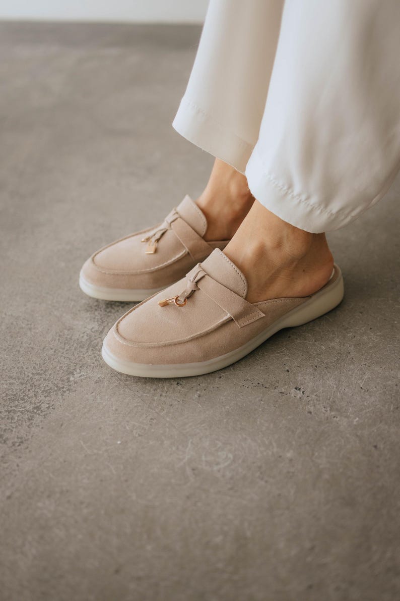 K&ouml;nnte beinhalten: Beige Wildleder-Slipper mit Quasten-Detail und einem kleinen goldfarbenen Akzent. Die Schuhe haben eine runde Spitze und eine niedrige, durchscheinende Sohle. Das Bild zeigt die Schuhe auf einer grauen Oberfl&auml;che.