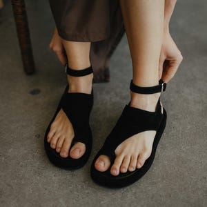 May include: Black sandals with a toe loop and ankle strap. The sandals have a thick sole and a minimalist design. The person is adjusting the ankle strap. The sandals are made of a soft material.