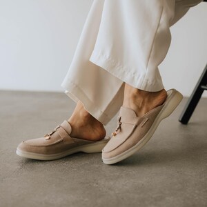 K&ouml;nnte beinhalten: Beige Wildleder-Slipper mit runder Spitze und einem kleinen goldfarbenen Akzent. Die Schuhe haben eine niedrige, wei&szlig;e Sohle und einen Riemen &uuml;ber der Oberseite. Das Bild zeigt die Schuhe auf einer grauen Oberfl&auml;che.