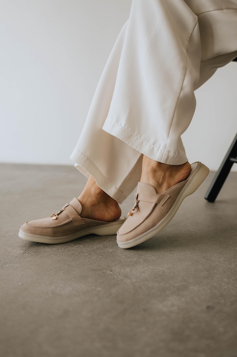 K&ouml;nnte beinhalten: Beige Wildleder-Slipper mit runder Spitze und einem kleinen goldfarbenen Akzent. Die Schuhe haben eine niedrige, wei&szlig;e Sohle und einen Riemen &uuml;ber der Oberseite. Das Bild zeigt die Schuhe auf einer grauen Oberfl&auml;che.