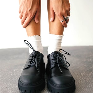May include: Black leather lace-up shoes with thick, lug soles. The shoes have a classic design with a rounded toe and visible stitching. White socks with a ruffled edge are visible above the shoes. The shoes are on a gray surface.