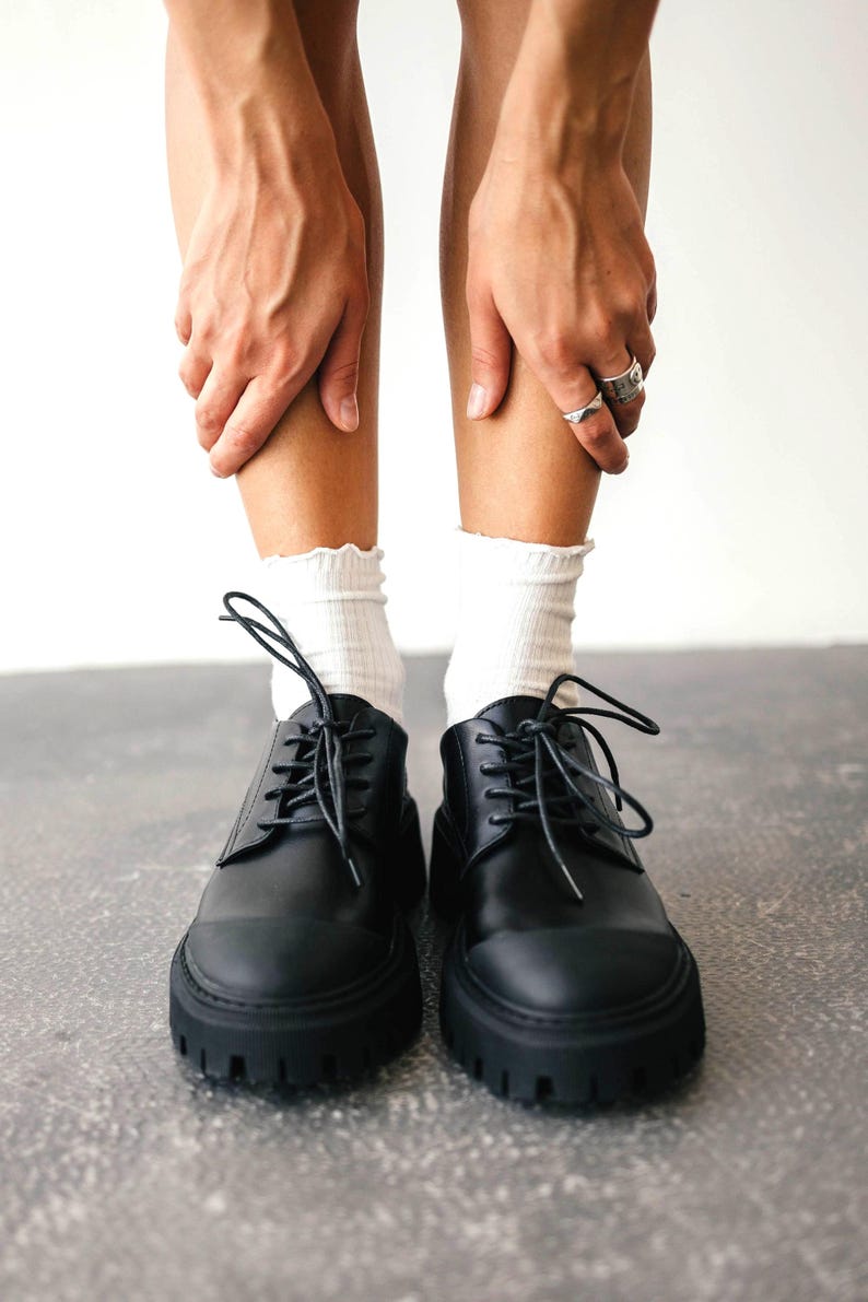 May include: Black leather lace-up shoes with thick, lug soles. The shoes have a classic design with a rounded toe and visible stitching. White socks with a ruffled edge are visible above the shoes. The shoes are on a gray surface.
