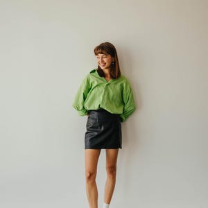 May include: A person wearing a lime green button-down shirt and a black leather mini skirt. The person is also wearing black shoes and white socks. The outfit is set against a plain white wall.