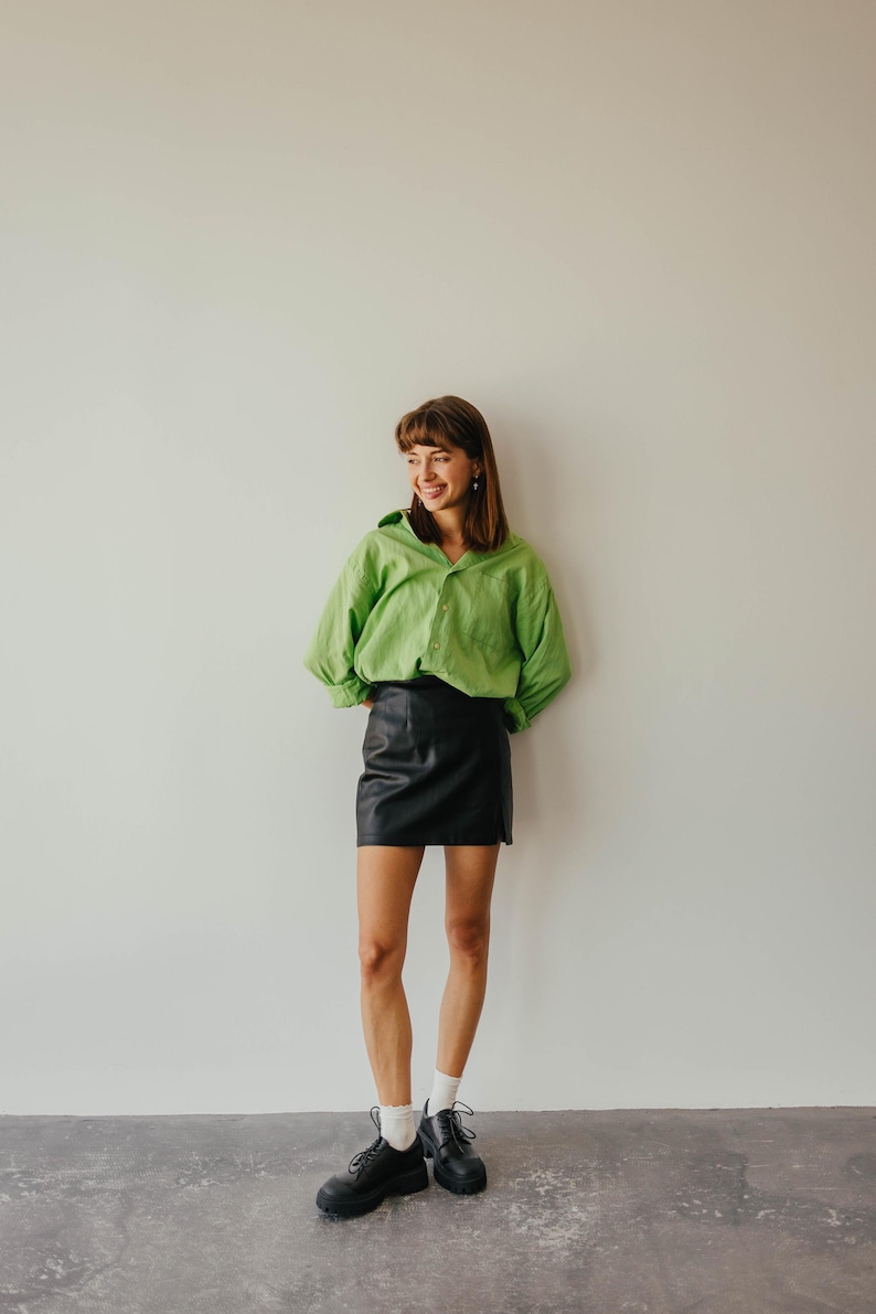May include: A person wearing a lime green button-down shirt and a black leather mini skirt. The person is also wearing black shoes and white socks. The outfit is set against a plain white wall.