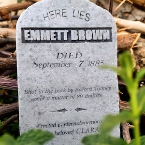Può includere: Una lapide grigia e consumata con l'iscrizione "HERE LIES EMMETT BROWN DIED September 7, 1885." Il testo descrive la causa della morte e una dedica a "beloved CLARA."