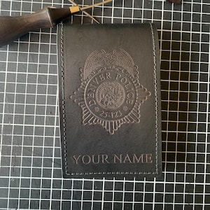 May include: Black leather Denver Police badge holder with embossed emblem and the text "YOUR NAME". Two leather working tools are placed above the badge holder on a black grid surface.
