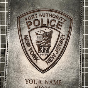 May include: Black leather badge holder with a brown embossed Port Authority Police emblem. The emblem includes an American flag, the numbers "37" and "9-11-01". The bottom of the holder is engraved with "YOUR NAME" and "#1234".