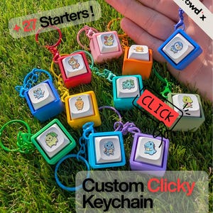 May include: A collection of colorful square keycap keychains, each featuring a small cartoon character illustration on the top surface. The keycaps are in various solid colors including blue, red, pink, orange, teal, yellow, green, and purple.