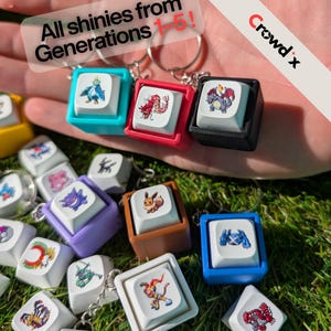 Könnte beinhalten: Eine Sammlung von quadratischen Tastenkappen-Schlüsselanhängern in verschiedenen Farben, die jeweils eine einzigartige Charakterillustration zeigen. Der Text "All shinies from Generations 1-5!" und das "Crowd x"-Logo sind oben zu sehen.