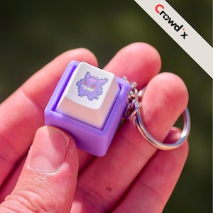 K&ouml;nnte beinhalten: Ein lila-wei&szlig;er Keycap-Schl&uuml;sselanh&auml;nger mit einem pixeligen Gengar-Design. Der Keycap ist in einem lila Rahmen eingefasst und an einem silbernen Schl&uuml;sselring befestigt. Der Text "Crowd x" ist oben rechts sichtbar.