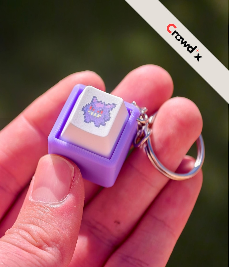 K&ouml;nnte beinhalten: Ein lila-wei&szlig;er Keycap-Schl&uuml;sselanh&auml;nger mit einem pixeligen Gengar-Design. Der Keycap ist in einem lila Rahmen eingefasst und an einem silbernen Schl&uuml;sselring befestigt. Der Text "Crowd x" ist oben rechts sichtbar.