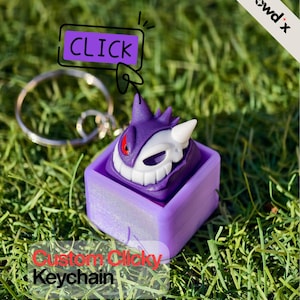 Peut inclure: Un porte-clés de personnage violet et blanc avec un porte-clés argenté. Le porte-clés est dans une base carrée violette. L'image comprend le texte "CLICK" et "Custom Clicky Keychain". L'arrière-plan est de l'herbe verte.