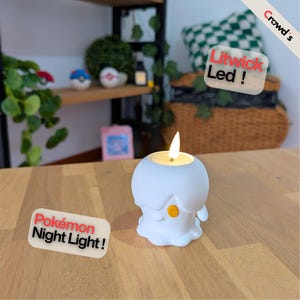 Puede incluir: Una luz nocturna LED Litwick blanca con forma de vela y un detalle amarillo, que presenta una bombilla brillante con forma de llama en la parte superior. El producto incluye etiquetas de texto Crowd's, Litwick Led ! y Pokémon Night Light !.
