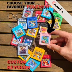 Op de afbeelding: Een verzameling miniatuur Game Boy cartridge sleutelhangers in diverse kleuren, waaronder blauw, rood, geel, roze, zwart en zilver. Elke sleutelhanger is voorzien van een label met klassieke videogame-illustraties en de tekst CHOOSE YOUR FAVORITE POKEMON.