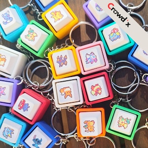 K&ouml;nnte beinhalten: Eine Sammlung bunter Schl&uuml;sselanh&auml;nger mit quadratischen Tasten, die pixelige Charakterdesigns zeigen. Die Tasten sind in leuchtenden Rahmen in Blau, Gr&uuml;n, Gelb, Rot, Orange und Lila gefasst und an silbernen Schl&uuml;sselringen befestigt. Der Text "Crowd x" ist sichtbar.