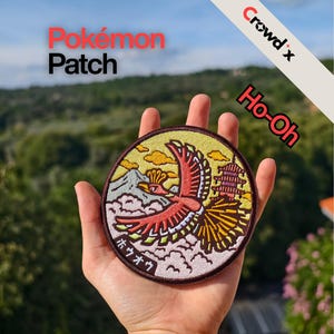 Puede incluir: Un parche Pokémon redondo con un pájaro rojo, amarillo y blanco volando sobre una montaña y nubes. El parche tiene el texto "Pokémon Patch" y "Ho-Oh". El parche se sostiene en una mano.