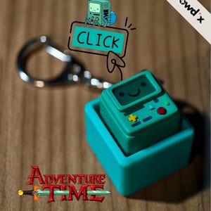 Könnte beinhalten: Ein türkisfarbener Adventure Time BMO-Schlüsselanhänger. Der Schlüsselanhänger hat eine türkisfarbene quadratische Basis mit einem kleineren BMO-Charakter oben. Das Wort "CLICK" befindet sich in einer Sprechblase über dem Schlüsselanhänger. Das Adventure Time-Logo befindet sich unten.