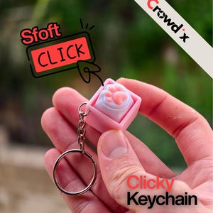 Könnte beinhalten: Ein pinkfarbener Clicky-Schlüsselanhänger mit Pfotenabdruck-Design. Der Schlüsselanhänger hat eine silberne Kette und einen Ring. Das Bild enthält den Text "Sfoft CLICK" und "Clicky Keychain". Das Crowd x-Logo befindet sich in der oberen rechten Ecke.