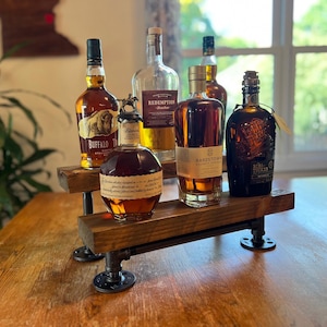 Könnte beinhalten: Ein hölzerner Spirituosen-Präsentationsständer mit schwarzen Rohrfüßen hält sechs Flaschen verschiedener Whiskeys. Die Flaschen haben unterschiedliche Formen und Größen, mit bernsteinfarbener Flüssigkeit im Inneren. Der Ständer steht auf einem Holztisch.