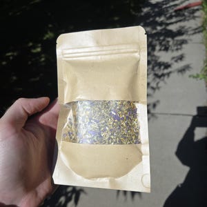 May include: A kraft paper pouch, partially open, filled with dried herbs and flowers. The contents are visible through a clear window. The pouch is held in a hand, with a green beaded bracelet visible on the wrist. The background is blurred.