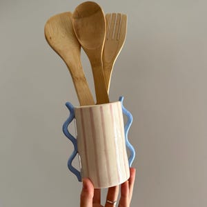 Può includere: Un portautensili in ceramica color crema con strisce verticali rosa e manici blu ondulati, contenente tre utensili da cucina in legno: un cucchiaio e una forchetta.