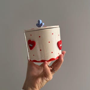 Può includere: Un vaso di ceramica con coperchio, tenuto in mano. Il vaso è bianco sporco con pois rossi e disegni a forma di cuore con occhi. Il coperchio ha una maniglia blu a forma di fiore e la base è rifinita in rosso. È un oggetto decorativo.
