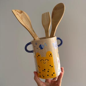 May include: A cylindrical ceramic utensil holder with two blue handles, featuring a speckled beige finish, a yellow spotted cat illustration, and small blue fish and moon motifs, containing three light brown wooden kitchen utensils.