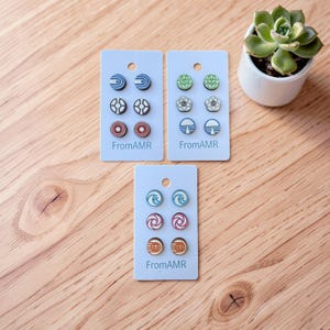 May include: Three sets of stud earrings on light blue cards. Each card features three pairs of earrings with various geometric and abstract designs in colors like blue, green, red, and brown. The text "FromAMR" is printed on each card.
