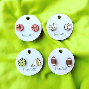 May include: Four pairs of sports-themed stud earrings on white cards. The earrings feature basketballs, volleyballs, pickleballs, and footballs. Each card has the text "FromAMR" printed on it. The background is a bright green fabric.
