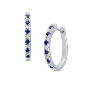 May include: A pair of silver hoop earrings with a row of small, round, dark blue gemstones and clear gemstones. The earrings are set against a white background.