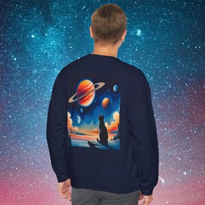 May include: Navy blue sweatshirt featuring a colorful illustration of a cat silhouetted against a cosmic landscape with planets and clouds. The artwork includes a large planet with rings and a vibrant sunset.
