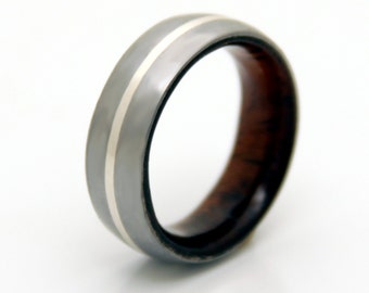 May include: A grey ring featuring a silver stripe and a dark wood interior. The ring is set against a white backdrop. The ring's design is simple and modern, suitable for everyday wear.