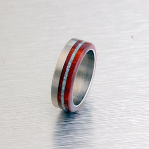 May include: A silver ring with a red and white wood inlay. The ring is made of titanium and has a polished finish.