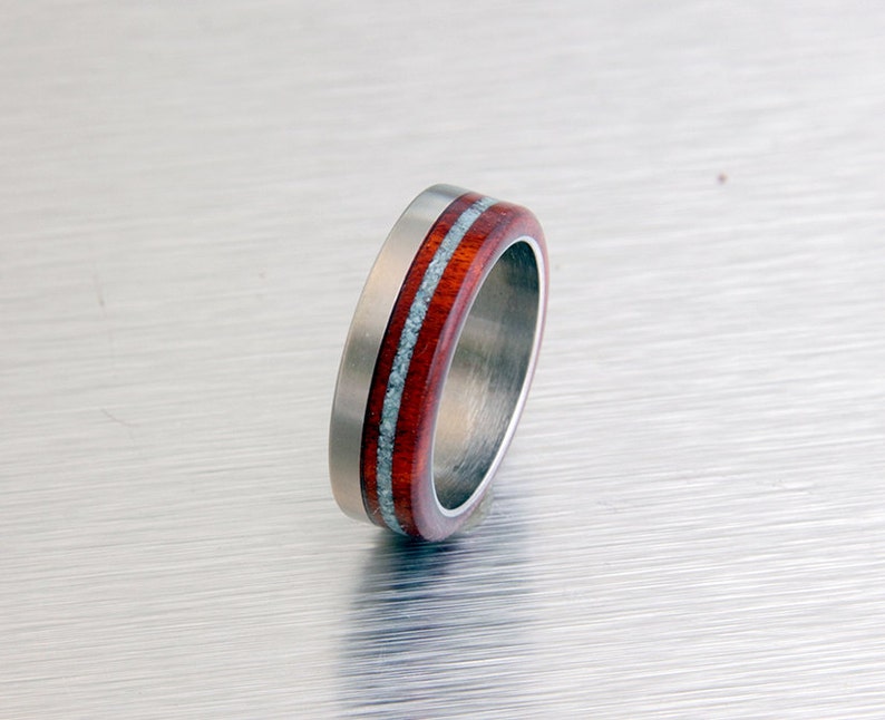 May include: A silver ring with a red and white wood inlay. The ring is made of titanium and has a polished finish.
