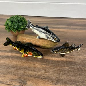 May include: Three decorative fish figurines. One is black and white, another is yellow, orange, and black, and the third is dark brown. They are arranged on a wooden surface with a small plant in the background.